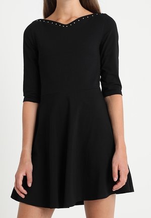 Femme portant une robe noire jusqu'aux genoux avec des manches trois-quarts et de petits clous argentés le long du large décolleté en V.