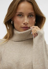 Bež pulover s turtleneck ovratnikom, ki ima rebrast ovratnik in manšete, izdelan iz mehkega in teksturiranega blaga ter sproščenega kroja.