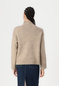 Beige stickad tröja med hög krage, ribbade mudd och kant. Mjuk textur, något löst passform, sedd bakifrån.