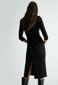 Abito aderente nero con maniche a tre quarti, spacco laterale sull'orlo e tessuto morbido. Modella di spalle con capelli lunghi e mossi.