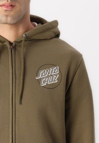 Olivno zelena zip-up kapuca iz mehkega materiala, ki ima krožni bel logo s tekstom "SANTA CRUZ" na levem prsnem delu.