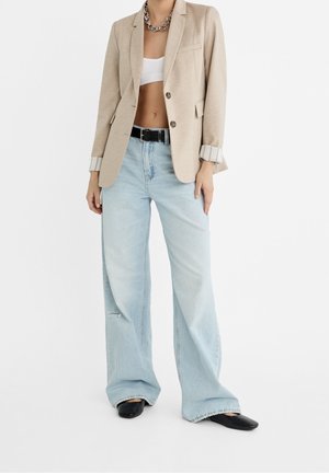 Blazer beige con un solo botón, combinado con jeans de pierna ancha de color azul claro con detalles desgastados y un cinturón negro. Zapatos negros de punta.