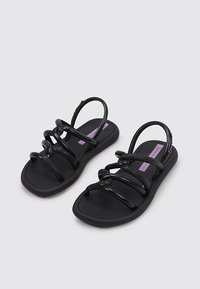 Sandalias negras con múltiples correas brillantes y una suela texturizada, con un logo morado en la plantilla. Diseño flexible para mayor comodidad.