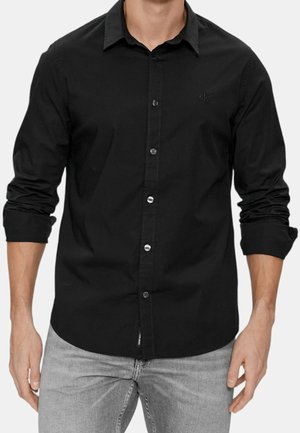 Mann trägt ein langärmliges schwarzes Hemd mit hochgekrempelten Ärmeln und hellgraue Jeans, steht mit entspannten Händen an den Seiten.