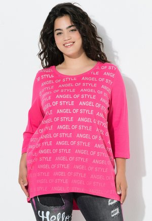 Violet lagu panjang dengan cetakan "ANGEL OF STYLE" dalam warna perak, dipadukan dengan jeans gelap yang robek. Bahan yang lembut dan potongan yang santai.