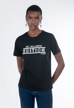 Persona joven con cabello corto que lleva pendientes en espiral, una camiseta negra que dice "Hola, soy una influencer de edición limitada" y jeans azules.