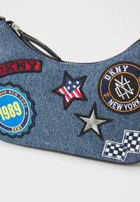 Jeanshandväska med olika broderade märken, inklusive stjärnor, logotyper och en rund badge. Dragkedja upptill.