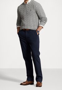 Šedý svetr s pleteným vzorem, námořnické přizpůsobené kalhoty, hnědé mokasíny s třásněmi, hodinky na zápěstí, viditelné manžety bílé košile, postoj ve stoje.