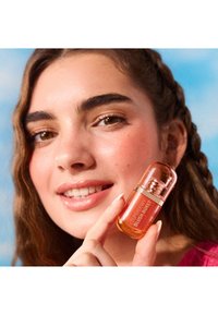 Small, round, amber-colored bottle with a transparent cap; labeled "SUPERDEWY BLUSH BURST." Background features a blue sky.