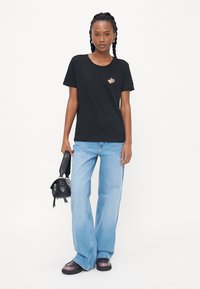 Svart bomull T-skjorte med korte ermer og et lite gullhjerte-grafikk, kombinert med lyse utsvingte jeans og svarte tilbehør.