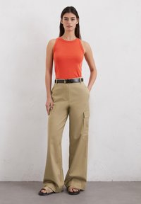 Top senza maniche rosso, pantaloni cargo larghi color khaki con tasche laterali, cintura nera e sandals flat neri. Sfondo neutro.
