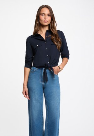 Femme aux longs cheveux bruns portant une chemise bleu marine boutonnée et nouée à la taille, ainsi qu’un jean bleu taille haute à jambes larges, debout avec une main dans la poche.