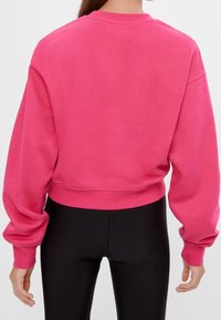 Roze cropped sweatshirt met lange mouwen, geribbelde kraag en boorden, zachte textuur, gedragen met zwarte leggings tegen een neutrale achtergrond.
