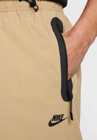 Shorts beiges en tissu léger avec une poche zippée noire et le logo Nike au bas. Comprend une taille ajustable avec cordon de serrage.