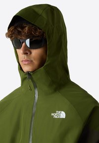 Giacca verde con cappuccio, realizzata in un tessuto leggero, con chiusura con zip e logo. Accompagnata da occhiali da sole scuri, lo stile è funzionale e casual.