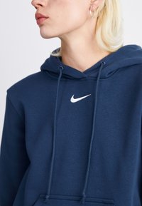 Tamsiai mėlynas Nike purkštukas su priekine kišenėle, raišteliais ir klasikiniu balto logotipo atvaizdu. Minkšta medžiaga su laisvu kirpimu ir veliūro kraštais.