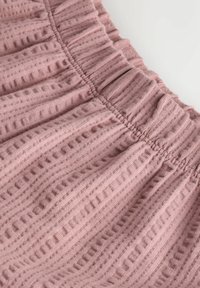 Weicher rosa gerippter Stoff mit einem gesammelten elastischen Bund und strukturierten vertikalen Streifen auf einem einfarbigen Hintergrund.