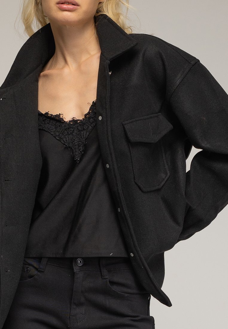 Chaqueta negra de gran tamaño con cierre de botones, dos bolsillos en el pecho y una textura suave. Se lleva sobre una blusa negra con encaje y pantalones negros.