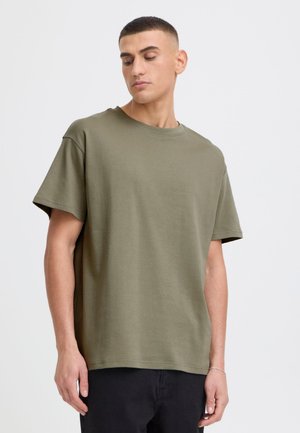 Ung mann med kort hår iført en olivengrønn oversized t-skjorte og svarte bukser, stående foran en ensfarget lys bakgrunn.