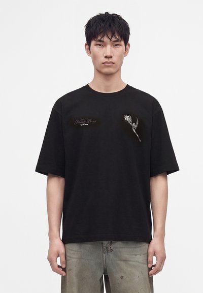 Νέος άνδρας φοράει μαύρο oversized T-shirt με μικρό διακοσμητικό κείμενο και γραφικά στο στήθος, συνδυασμένο με φθαρμένο γκρι παντελόνι.