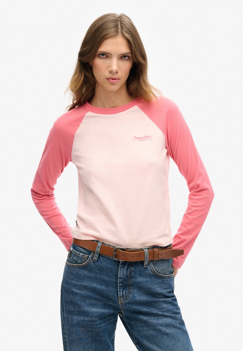 Jonge vrouw met lang haar, gekleed in een roze en lichtroze raglan shirt met lange mouwen, blauwe spijkerbroek en een bruine riem, staand met haar handen achter haar rug.