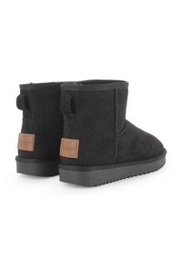 Botas de ante negras hasta el tobillo con suela de goma plana, que presentan una tira de agarre y un acento de cuero marrón claro en la parte trasera. Textura suave y diseño sencillo.