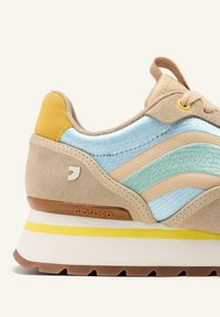 Zapatilla de ante beige con detalles de tela azul claro, tacón amarillo y entresuela blanca con una franja amarilla. Logo de Gioseppo visible.