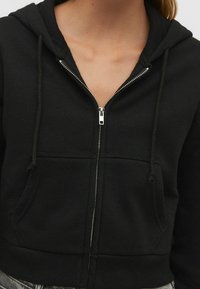 Hoodie zippé noir en tissu doux. Doté d'une capuche à cordon de serrage, de poches avant et de détails en métal argenté sur la fermeture éclair.