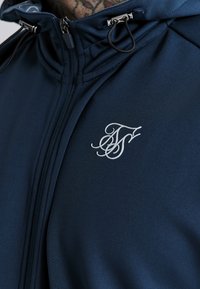 Tamsiai mėlyna užtrauktuku užsegama striukė, pagaminta iš lygios medžiagos, su sidabriniu užtrauktuku, reguliuojamais raišteliais ir balta inicialų logo ant krūtinės.