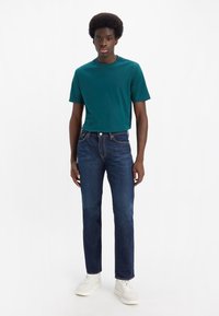 Teal t-skjorte med korte ermer, blå jeans og hvite sneakers. T-skjorten har rund hals, og jeansene har rette ben med synlige sømdetaljer.
