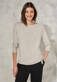 Maglietta grigia chiara a maniche lunghe con scollatura rotonda ampia, sottili accenti testurizzati sulle maniche e un dettaglio annodato all'orlo. Pantaloni neri.