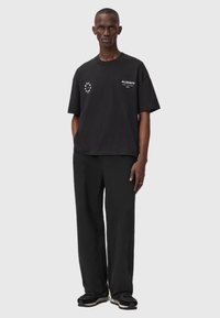 Must tume T-särk, millel on graafiline logo rinnal ja õlal. Sobitatud mustade avarate pükstega ja mustade tossudega.