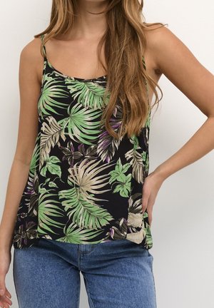 Nainen pukeutunut mustaan hihattomaan paitaan, jossa on vihreä ja beige trooppinen lehtikuvio, sekä sinisiin farkkuihin, käsivarsi lepää lantiolla.