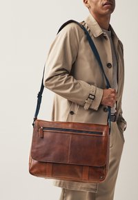 Geantă messenger din piele maro cu finisaj texturat, curea albastră reglabilă, buzunar frontal cu fermoar și închidere cu clapetă.
