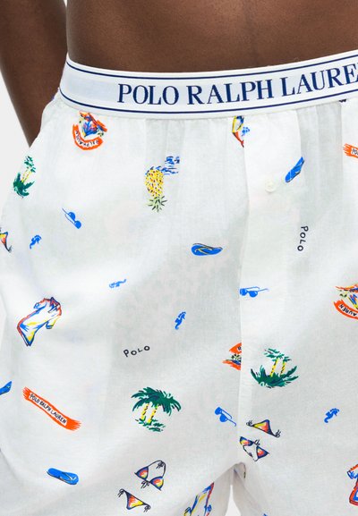 Κοντινό πλάνο λευκών μποξεράκια με πολύχρωμα τροπικά σχέδια και ζώνη μέσης "Polo Ralph Lauren" σε άτομο με σκουρόχρωμο δέρμα.