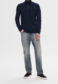 Man som bär en marinblå tröja med halvmatch, ljusa urtvättade jeans och svarta slip-on sneakers med vita sulor, står med en hand i fickan.