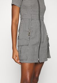 Lyhythihainen, vartalonmyötäinen mustavalkoinen koirankynttiläkuvioinen mekko, jossa on etuvetoketju ja sivuilla vetoketjulliset taskut. Sileä pinta.