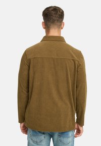 Mand vender ryggen iført en olivengrøn fløjljakke og lyseblå denimjeans foran en ensfarvet hvid baggrund.