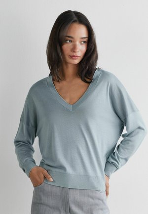 Jeune femme aux cheveux bruns mi-longs portant un pull bleu clair à col en V et un pantalon gris, debout les mains dans les poches.