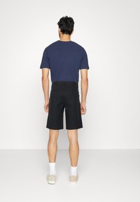 Dunkle Shorts mit lässiger Passform, fransigem Saum und Gesäßtaschen. Kombiniert mit einem marineblauen T-Shirt und beigen Sneakers. Sauberes, minimales Design.