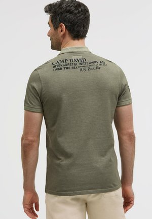 Mann mit dem Rücken zur Kamera, trägt ein olivgrünes Poloshirt mit schwarzem Text auf dem oberen Rücken und beige Hose.
