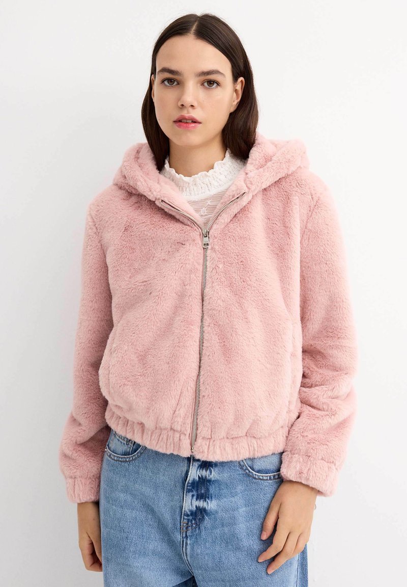 Chaqueta de piel sintética de color rosa claro, con capucha, cierre de cremallera y dobladillo elástico. Presentada sobre una blusa blanca con volantes y un cuello, y jeans de denim.