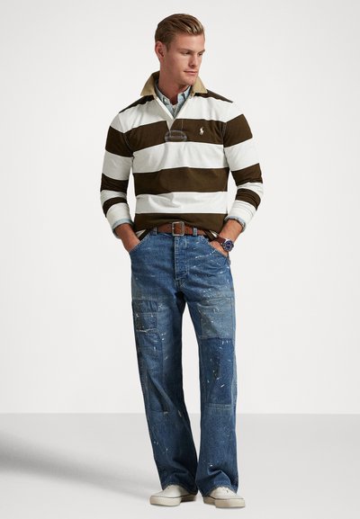 Vīrietis, kurš valkā brūnu un baltu svītrainu regbija krekli, plankumainas džinsas ar krāsas traipiem, baltas sporta kurpes un rokassprādzi, stāv ar rokām kabatās.