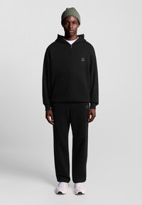 Felpa nera con zip, tasche a marsupio e logo, abbinata a pantaloni neri coordinati. Il modello indossa un berretto verde chiaro e sneakers bianche.