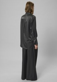 Kvinde der står vender væk iført en løs sort satin skjorte og brede sorte bukser mod en neutral baggrund.