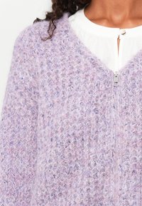 Lila gestrickte Strickjacke mit Reißverschluss. Das strukturierte Garn zeigt eine Mischung aus helleren und dunkleren Lilatönen und hat ein weiches, flauschiges Aussehen.