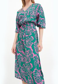 Mujer que lleva un vestido midi verde con estampado floral en rosa, escote en V, mangas abullonadas y cintura elástica con un cinturón de tela a juego.