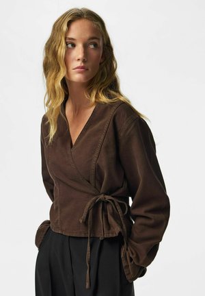 Ung kvinde med bølget blondt hår iført en brun slå-om-bluse med lange ærmer og sorte bukser, der kigger mod venstre.
