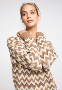 Pulover tricotat cu un model în zigzag în nuanțe de bej, crem și maro. Mansetele ribbate și gulerul rotund subliniază designul. Textura moale este vizibilă.
