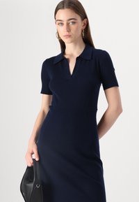 Abito a coste blu navy con maniche corte, colletto e design aderente. La modella tiene una borsa nera, mostrando una texture liscia e una silhouette aderente.